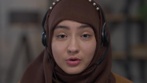 Retrato en la cabeza de una joven confiada mujer de Oriente Medio con auriculares y hijab hablando mirando a la cámara. Primer plano de chat de vídeo POV de consultor profesional de negocios experto en la oficina en casa. — Vídeo de stock