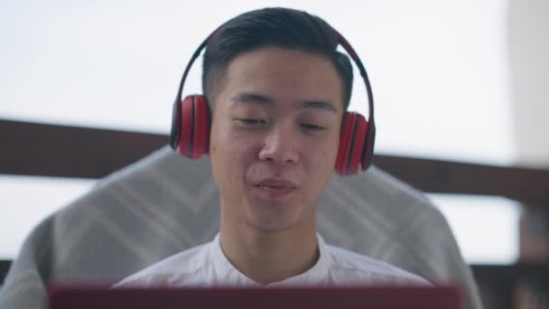 Gros plan du visage souriant bel homme asiatique dans les écouteurs en regardant le chat web ordinateur portable parler. Headshot de joyeux gestionnaire chinois heureux confiant en utilisant conférence virtuelle assis dans le bureau à la maison sur le pont — Video