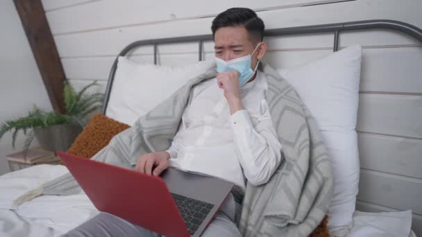 Épuisé malade asiatique jeune homme couché sur le lit tousser surfer sur Internet sur ordinateur portable. Portrait d'un gestionnaire chinois malade en masque facial travaillant en ligne sur l'auto-isolement lors d'une pandémie de coronavirus. — Video