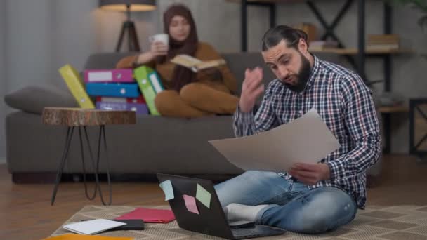Στοχαστική σοβαρή όμορφος άνθρωπος της Μέσης Ανατολής ανάλυση επιχειρηματικό γράφημα κάθεται στο σπίτι γραφείο σύζυγος ανάγνωση στο παρασκήνιο. Πορτρέτο του εστιασμένου επαγγελματία που εργάζονται σε απευθείας σύνδεση χρησιμοποιώντας συνομιλία βίντεο στο φορητό υπολογιστή. — Αρχείο Βίντεο