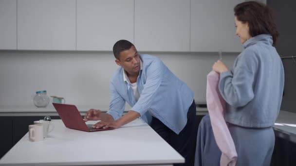 Pandangan samping terhadap pria Afrika-Amerika yang terkonsentrasi serius mengetik di papan ketik laptop dalam gerakan lambat sebagai wanita Kaukasia yang mengalihkan perhatian suami dari pekerjaan rumah tangga yang jauh di dapur. Konsep ketidaknyamanan — Stok Video