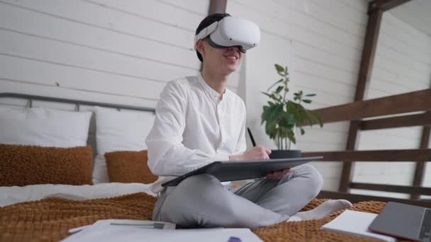 Visão de ângulo lateral do designer asiático criativo talentoso positivo no desenho de fone de ouvido VR no tablet gráfico sentado na cama dentro de casa. Sorrindo jovem chinês trabalhando em realidade aumentada. — Vídeo de Stock