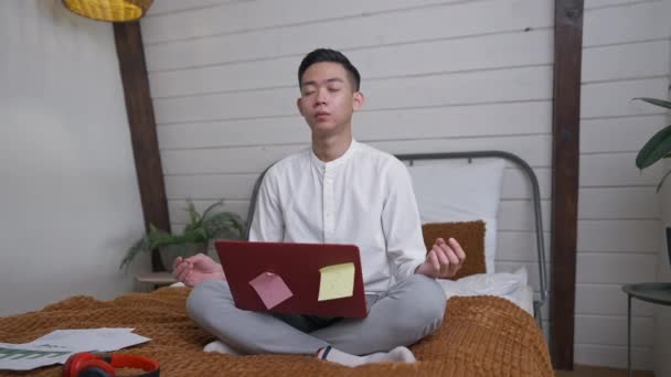 Portrait of calm Chinese young man sitting in yoga lotus pose with closed eyes and laptop on legs. Wide shot of handsome Asian guy having break working online in home office bedroom indoors. — Stock Video