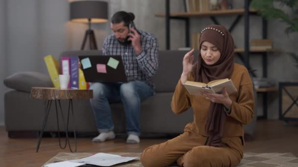 Irriterad ung smal vacker muslimsk kvinna skriker på nervös make argumentera på telefon i bakgrunden. Missnöjd Mellanösterns fru i hijab distraherad från att läsa med mannen som arbetar på hemmakontoret. — Stockvideo