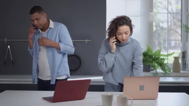 Ocupado hombre afroamericano y mujer caucásica hablando por teléfono trabajando en línea en la cocina en casa. Expertos profesionales serios en el bloqueo del coronavirus. Nuevo estilo de vida normal. — Vídeo de stock