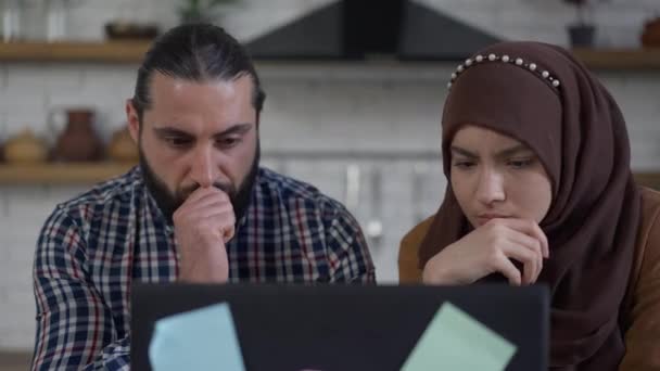 Pensativo confuso marido y mujer de Oriente Medio hablando de la planificación de la estrategia de inicio en la oficina en el interior. Retrato del hombre y la mujer concentrados analizando el mercado en línea utilizando el ordenador portátil en la cocina. — Vídeos de Stock