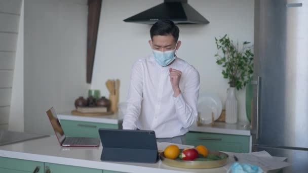 Ung kinesisk man står i köket surfa Internet på surfplatta och hosta. Porträtt av sjuk asiatisk kille i Covid-19 ansiktsmask med symtom på coronavirus sjukdom arbetar online på hemmakontoret. — Stockvideo