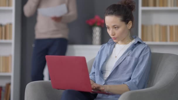 Upptagen vit ung kvinna skriva på laptop tangentbord arbetar på hemmakontoret ser tillbaka på oigenkännlig afroamerikansk man distraherande prata i telefonen i bakgrunden. Begreppet olägenhet. — Stockvideo