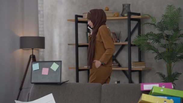 Wide shot of serious confident professional Middle Eastern young woman in hijab walking in living room talking on the phone and checking e-mail on laptop. Remote working in home office. — 비디오