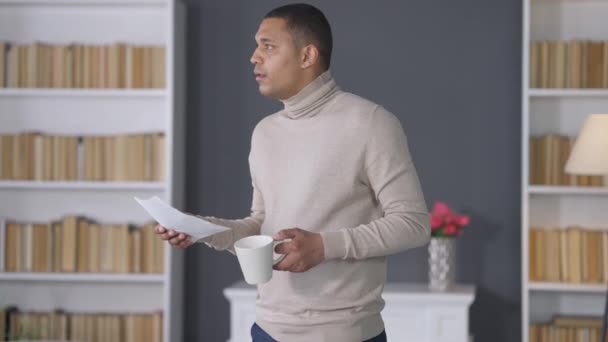 Thoughtful young African American man with coffee cup thinking analyzing documents turning to camera smiling. Portrait of confident positive handsome manager working in home office indoors. — 비디오