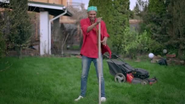 Breed shot van vrolijke jongeman harken en nabootsen gitaar spelen op hark zingen en glimlachen. Portret van grappige vrolijke knappe Afro-Amerikaanse man die plezier heeft in de tuin op zomerdag. — Stockvideo