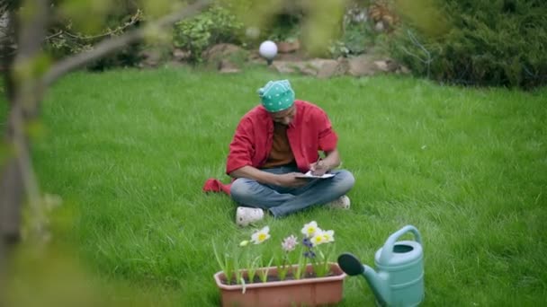 Großaufnahme eines zufriedenen afroamerikanischen Gärtners, der auf grünem Rasen im Hinterhof sitzt und Tagebuch schreibt. Porträt eines glücklich lächelnden, gutaussehenden jungen Mannes, der in der Pause im Garten arbeitet. Floristik und Lebensstil — Stockvideo