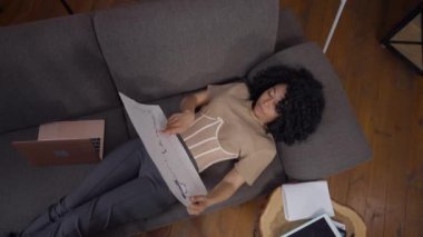 Ciddi profesyonel iş analistinin merkez ofisteki kanepede uzanan grafiği incelerken geniş açılı görüntüsü. Güzel, sıska Afrikalı Amerikalı genç bir kadın kapalı alanda strateji analiz ediyor..