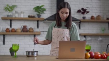Sırıtan ince Asyalı bir kadının yemek pişirme kabında laptopa bakarken orta pozu. Önlüklü güzel, pozitif şefin portresi. Mutfakta ev yapımı lezzetli yemekler hazırlıyor..