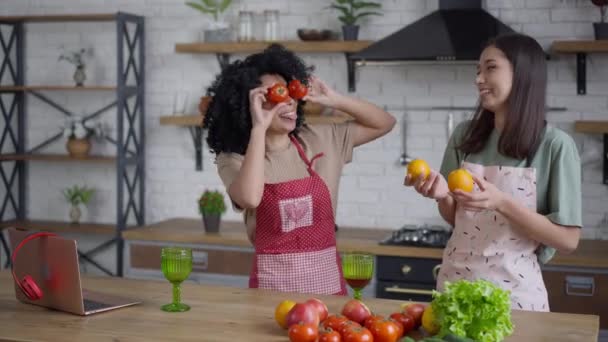 Ontspannen vrolijke vrienden die plezier hebben met tomaat en sinaasappel op grimmige ogen. Lachende vrolijke Afro-Amerikaanse en Aziatische vrouwen maken gezichten koken in de keuken thuis. Genot en culinair. — Stockvideo