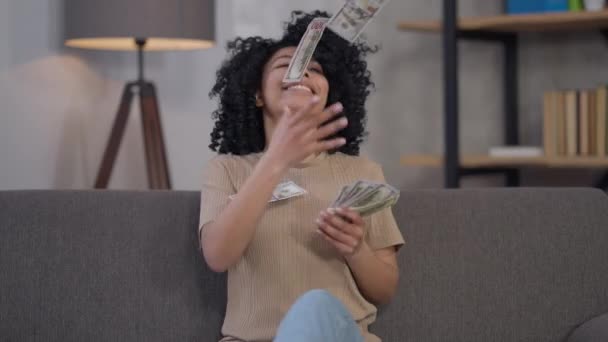 Счастливая богатая афро-американская молодая женщина, разбрасывающая деньги, сидя дома в офисе. Средний снимок портрет богатого веселого красивого стартапера фрилансера, радующегося успеху бизнес-идеи. — стоковое видео