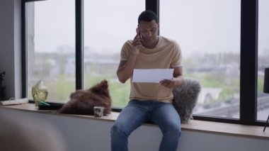 Endişeli bir Afrikalı Amerikalı iş grafiğini analiz ediyor. Ofisteki pencere eşiğinde oturuyor. Endişeli genç müdürün portresi. İçerde uzaktan planlama stratejisi..