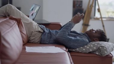 Kanepede baş aşağı uzanmış akıllı telefondan mesajlaşan genç adamın yan görüntüsü. Yakışıklı Afro-Amerikan özgüvenli girişimci samimi ofisten mesaj atıyor. Uzak çalışma konsepti.