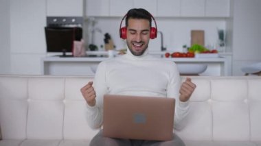 Kulaklıklı akıllı, tatmin olmuş Orta Doğulu bir adam koltukta dizüstü bilgisayarla oturmuş zafer jestleri yapıyor. Heyecanlı genç ve yakışıklı iş adamının portresi ev ofisinde başarılı bir başlangıç yapıyor..