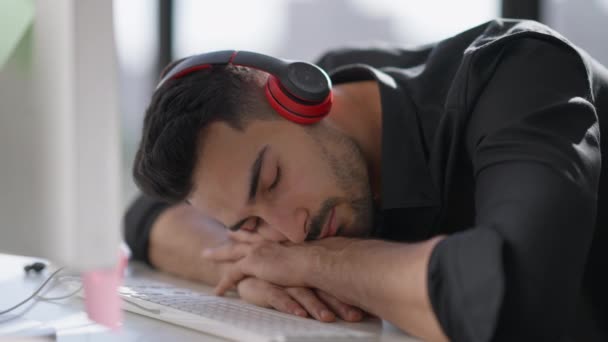 Κοντινό πλάνο πορτρέτο του εξαντλημένου νεαρού άντρα της Μέσης Ανατολής με ακουστικά που κοιμάται στο τραπέζι στο πληκτρολόγιο του υπολογιστή την ηλιόλουστη μέρα. Κουρασμένος εργατικός τύπος κοιμάται στο γραφείο στο σπίτι κατά τη διάρκεια της εργασίας. — Αρχείο Βίντεο