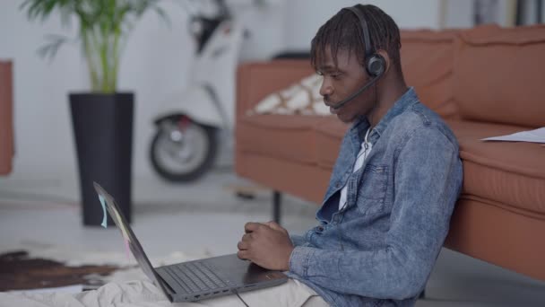 Vista lateral del joven en los auriculares hablando en el chat de vídeo en el ordenador portátil que explica la idea de negocio que muestra el gráfico. Confianza afroamericana startuper conferencia en línea desde su oficina en el hogar. — Vídeos de Stock