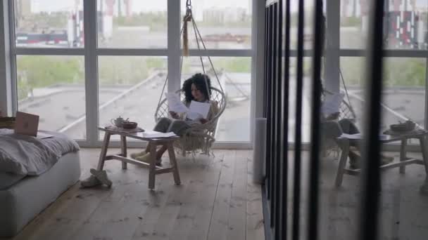 Vue d'ensemble extrême de la femme afro-américaine assise sur une chaise suspendue analysant la paperasse. Portrait de jeune manager confiant travaillant dans la chambre à coucher de bureau à domicile à l'intérieur. Concept freelance. — Video