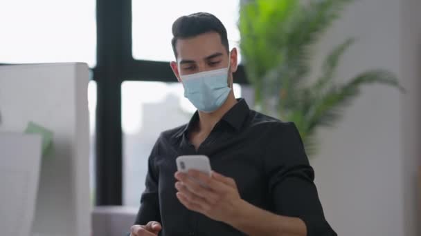 Cheerful young man in Covid-19 face mask taking selfie on smartphone in home office on sunny day. Portrait of smiling joyful positive Middle Eastern manager enjoying remote working. Lifestyle. — Stock Video