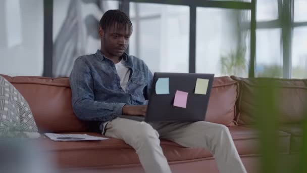 Portret van de jonge zelfverzekerde manager surfen op internet op laptop het analyseren van papierwerk zittend op een gezellige bank in huis kantoor woonkamer. Ernstig geconcentreerde Afro-Amerikaanse man die op afstand werkt. — Stockvideo