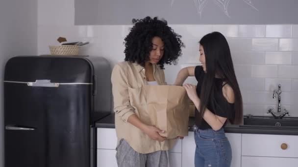 Femei tinere râzând despachetând cumpărăturile de alimente în bucătărie în interior. Colegii de cameră asiatici și afro-americani zâmbesc pozitiv gătind alimente sănătoase în interior vorbind. Conceptul de stil de viață . — Videoclip de stoc