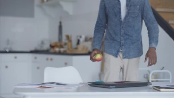 Joven hombre profesional caminando desde la cocina con manzana a la mesa en la oficina en casa sentado y la apertura de la computadora portátil. Retrato de un empleado afroamericano seguro que trabaja en línea por la mañana. — Vídeos de Stock