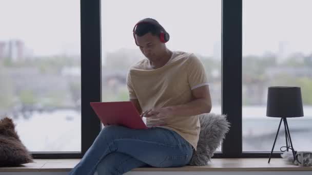 Junger Mann mit Kopfhörern winkt in Zeitlupe in die Laptop-Webkamera und unterhält sich auf der Fensterbank im Home Office. Porträt eines gutaussehenden Afroamerikaners bei Online-Konferenzen in Innenräumen. — Stockvideo