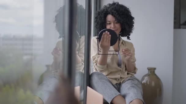 Plan large d'heureuse femme afro-américaine souriante admirant le reflet dans le miroir de la main. Portrait de jeune femme confiante à l'intérieur à la maison. Beauté et féminité. — Video