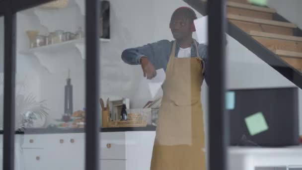 Радостный молодой афроамериканец танцует с бумажной работой на домашней кухне. Портрет веселого красивого парня в фартуке, веселящегося, радующегося успеху стартапа. Стиль жизни и радость. — стоковое видео