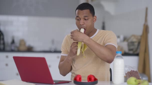 Joven afroamericano pelando y comiendo plátano sentado en la mesa de la cocina en la oficina del hogar. Retrato de plano medio de empleado enfocado trabajando en línea en el ordenador portátil escribiendo en el teclado almorzando. — Vídeo de stock