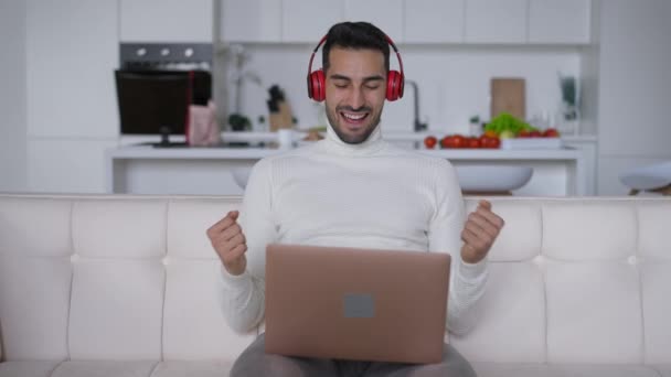 Spokojenej inteligentní muž ze Středního východu ve sluchátkách dělá vítězný gesto, sedí na gauči s laptopem. Portrét vzrušeného mladého pohledného podnikatele radující se ze startovního úspěchu v domácí kanceláři. — Stock video