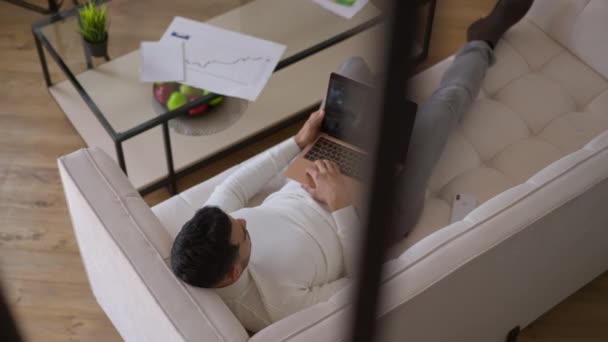High angle view of young Middle Eastern man using virtual conference on laptop lying on couch in home office. Professional colleagues business partners discussing strategy idea online. New normal. — Stock Video