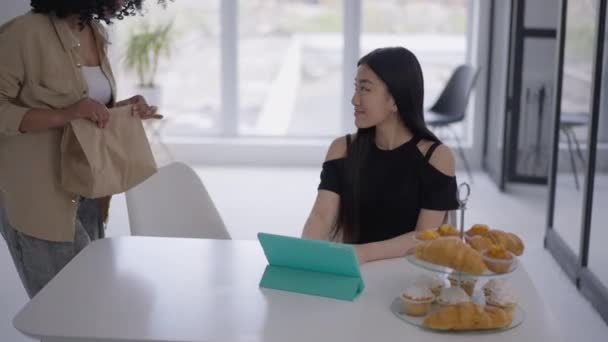 Mujer joven afroamericana que entra en la sala de estar trayendo comida para llevar para un amigo asiático que trabaja en la oficina en casa. Retrato de dos mujeres positivas sentadas a la mesa en el interior sonriendo y hablando. — Vídeos de Stock