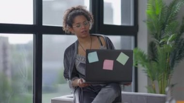 Kendine güveni tam genç bir kadının portresi. Uyku tulumu ve gözlüklü. Koltukta oturmuş dizüstü bilgisayarla düşünüyor. Afro-Amerikalı iş kadını sabahları e-postaları merkez ofiste kontrol ediyor..