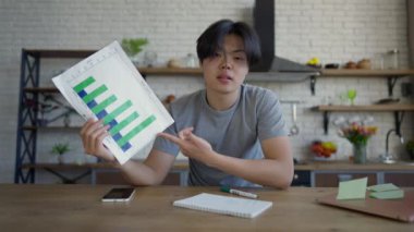 İş grafiğine işaret eden pozitif özgüvenli Asyalı uzman görüntülü sohbet POV 'u konuşuyor ve gülümsüyor. Genç adam ofisten çevrimiçi konferans yapıyor. Mutfak masasında oturuyor..