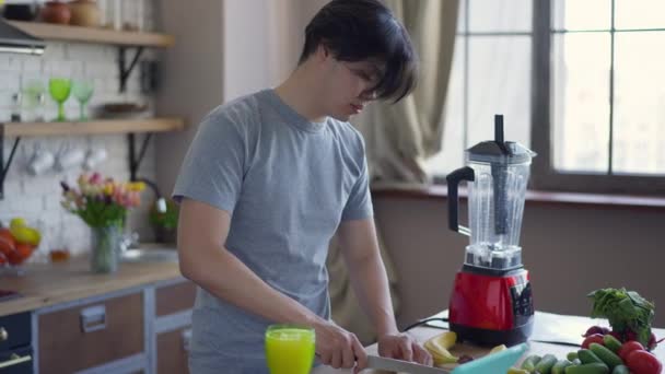 Mutfaktaki tezgahın üstünde bıçakla muz doğrayan Asyalı genç bir adamın yan görüntüsü. Rahatlamış mutlu milenyum portresi evde sağlıklı meyve suyu için meyve kesme sabahları. — Stok video