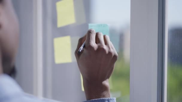 Close-up van Afro-Amerikaanse jonge mannelijke handschrift met pen op plakkerige notities op het raam. Onherkenbare man handschrift zakelijke ideeën in huis kantoor binnen op zonnige dag. — Stockvideo