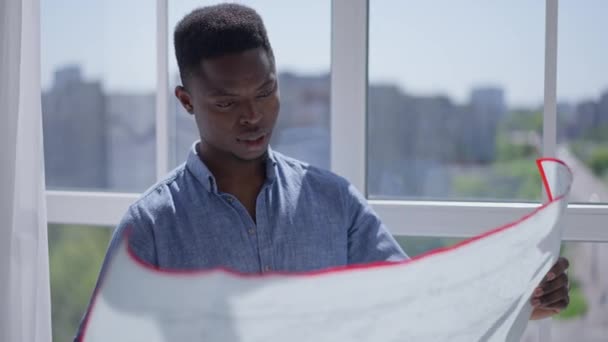 Fiatal kreatív okos kertész építész, aki az otthoni iroda ablakában álló tervrajzot vizsgálja. Portré magabiztos afro-amerikai tehetséges férfi elemzése projekt beltéri napsütéses napon. — Stock videók