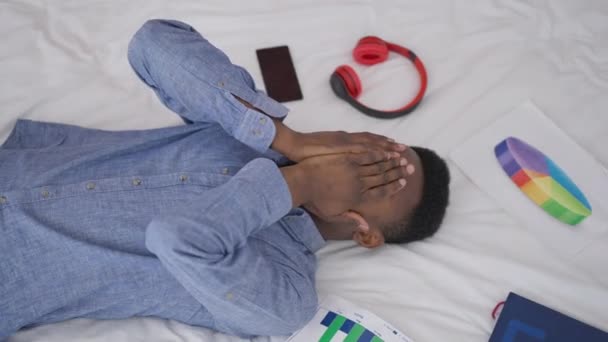 Widok z góry wyczerpany Afroamerykanin przedsiębiorca leżący na łóżku z wykresami i smartfon dotykając wzdychanie twarzy i myślenia. Portret zmęczonego zaniepokojonego człowieka myślącego o zarządzaniu kryzysowym. — Wideo stockowe