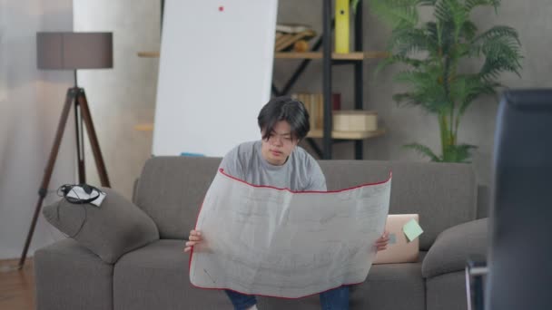 Wide shot of young Asian man examining landscape blueprint sitting on couch in home office. Portrait of thoughtful talented landscaper planning project in comfortable living room indoors. — Vídeos de Stock