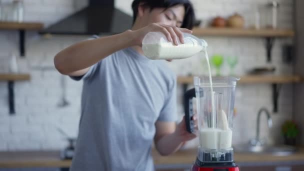 Câmera se aproxima em câmera lenta para leite derramando no liquidificador com banana picada. Jovem homem asiático preparando coquetel de frutas saudáveis na cozinha em casa pela manhã. — Vídeo de Stock