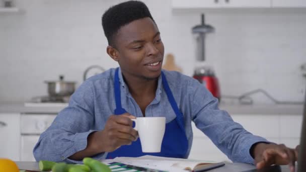 Freelancer jovem feliz digitando no teclado do laptop segurando xícara de café da manhã. Retrato de jovem afro-americano positivo sentado à mesa da cozinha em casa mensagens de escritório surf Internet. — Vídeo de Stock