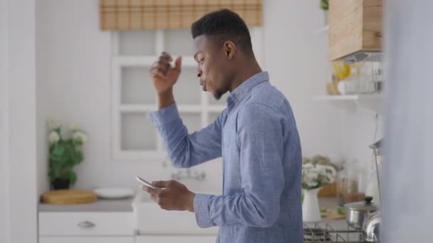 Pohled na vzrušeného mladého Afroameričana, jak dělá vítězné gesto a kontroluje e-mail na smartphonu v kuchyni. Šťastný spokojený úspěšný manažer v domácí kanceláři radující se z úspěchu. — Stock video