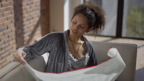 Young woman stretching neck examining architectural blueprint in sunny home office in the morning. Portrait of beautiful African American architect suffering pain from sedentary lifestyle. — Vídeo de stock