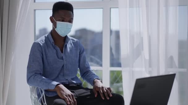 Jongeman in Coronavirus gezichtsmasker zittend in zonnig kantoor thuis online op laptop. Portret van een geconcentreerde Afro-Amerikaanse zelfverzekerde werknemer met behulp van videochat op Covid-19 lockdown. — Stockvideo