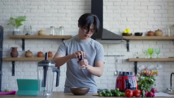 Middle shot of young Asian man salting healthful spring salad in kitchen in the morning. Portrait of confident handsome guy cooking healthy food indoors at home. Domestic routine and lifestyle. — Vídeos de Stock